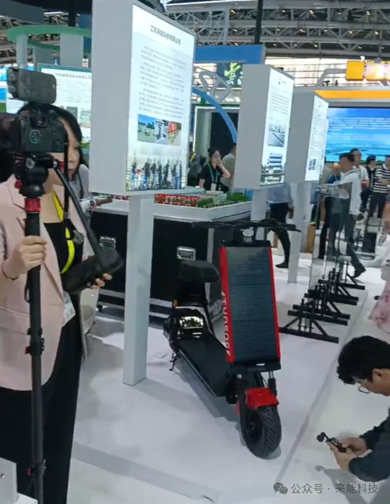 Female journalist filming Tuneng T87S Pro solar electric scooter with gimbal camera while photographer captures images from low angle at Laineng Technology exhibition booth during 19th Xi'an Silk Road Expo media coverage session