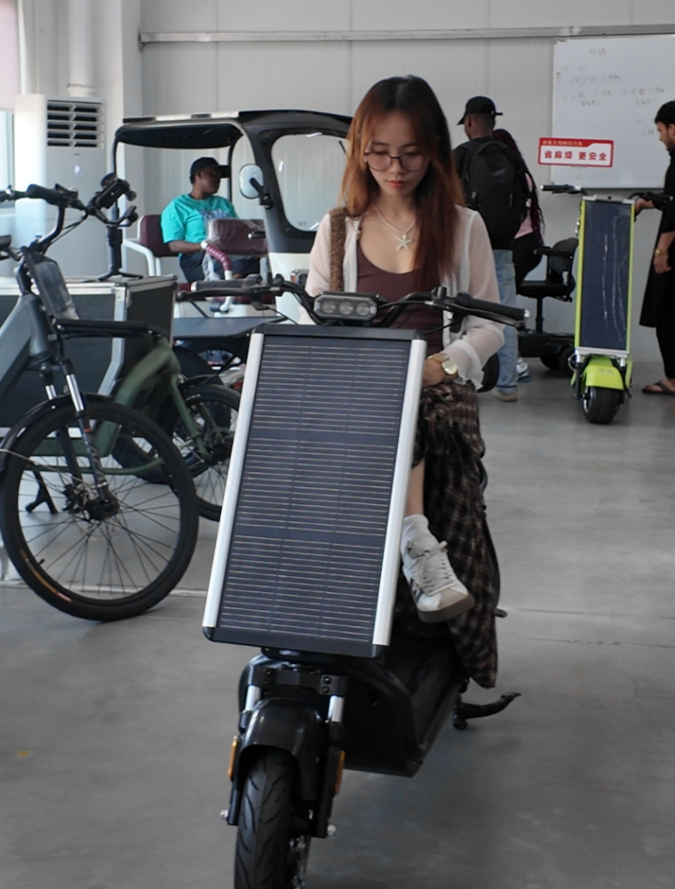 Female influencer seated on black Laineng solar kick scooter with large integrated photovoltaic charging panel at indoor brand center test area with solar e-bike and solar three-wheeler displayed in background
