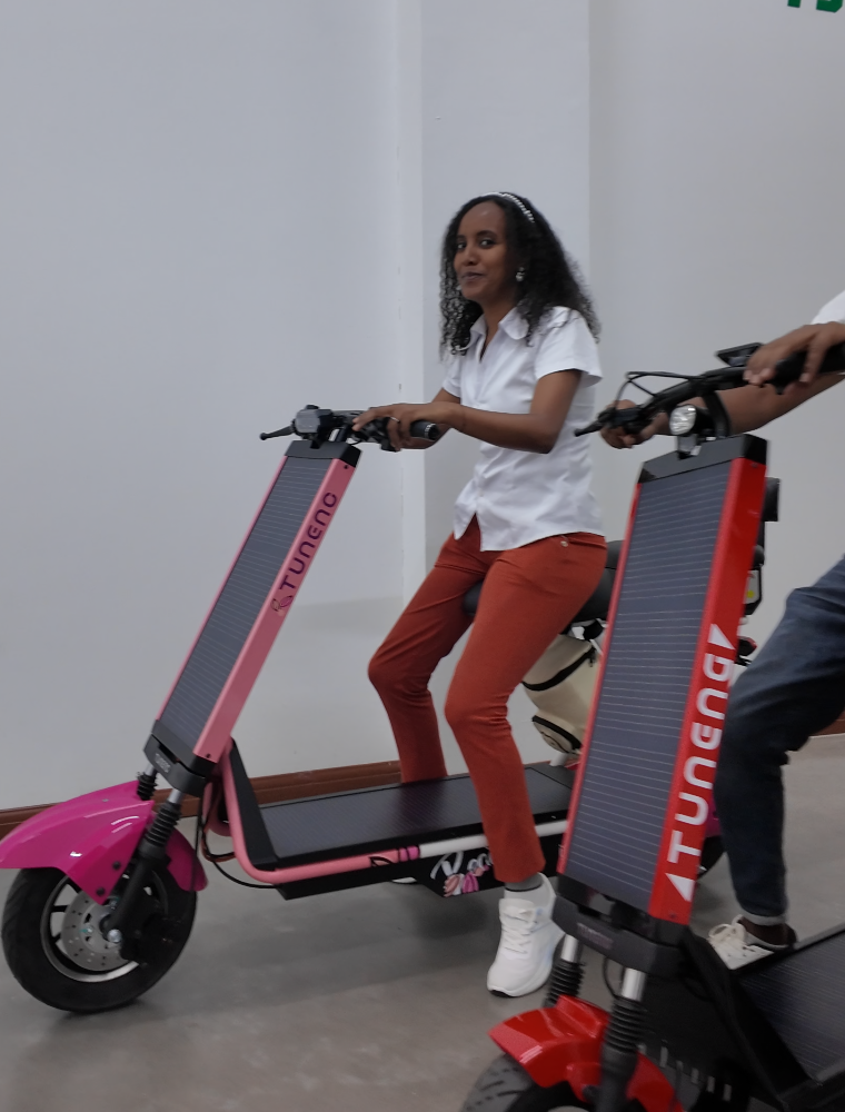 African influencer riding pink Laineng solar electric scooter alongside red model during group test ride session at brand center showcasing multiple color options and integrated PV panel design